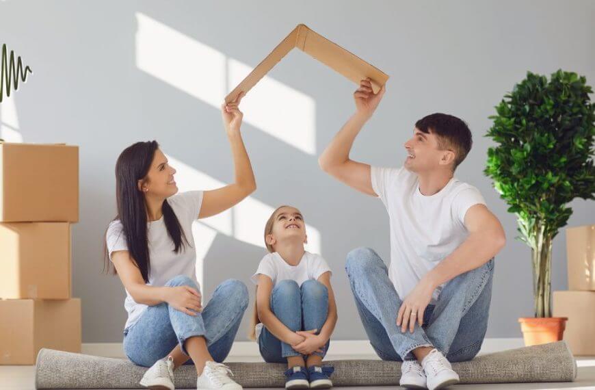 Smiling family sitting on a rolled-up rug in a new home surrounded by boxes, creating a supportive space for their hard-of-hearing child.