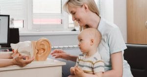 A mother, cradling her infant, attentively sits across from a dedicated hearing specialist. They're situated in what seems to be a state-of-the-art medical office. The specialist is passionately demonstrating an intricate anatomical model of the ear, showcasing its complexities and is getting ready for hearing screenings of the infant. This heartfelt interaction epitomizes our mission at the foundation—to enlighten and empower hard of hearing youth and their families with essential knowledge and support, fostering a brighter future for all.
