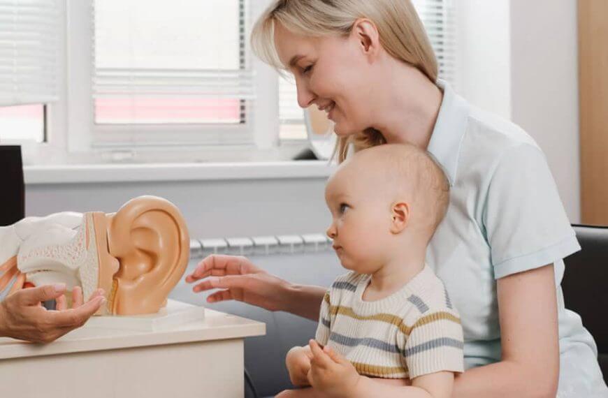 A mother, cradling her infant, attentively sits across from a dedicated hearing specialist. They're situated in what seems to be a state-of-the-art medical office. The specialist is passionately demonstrating an intricate anatomical model of the ear, showcasing its complexities and is getting ready for hearing screenings of the infant. This heartfelt interaction epitomizes our mission at the foundation—to enlighten and empower hard of hearing youth and their families with essential knowledge and support, fostering a brighter future for all.